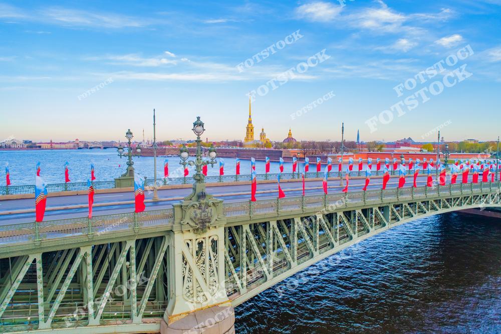 На фотографии изображён Троицкий мост, один из центральных мостов Санкт-Петербурга, украшенный в честь праздника. На заднем плане возвышается Петропавловская крепость с её знаменитым собором и золотым шпилем.